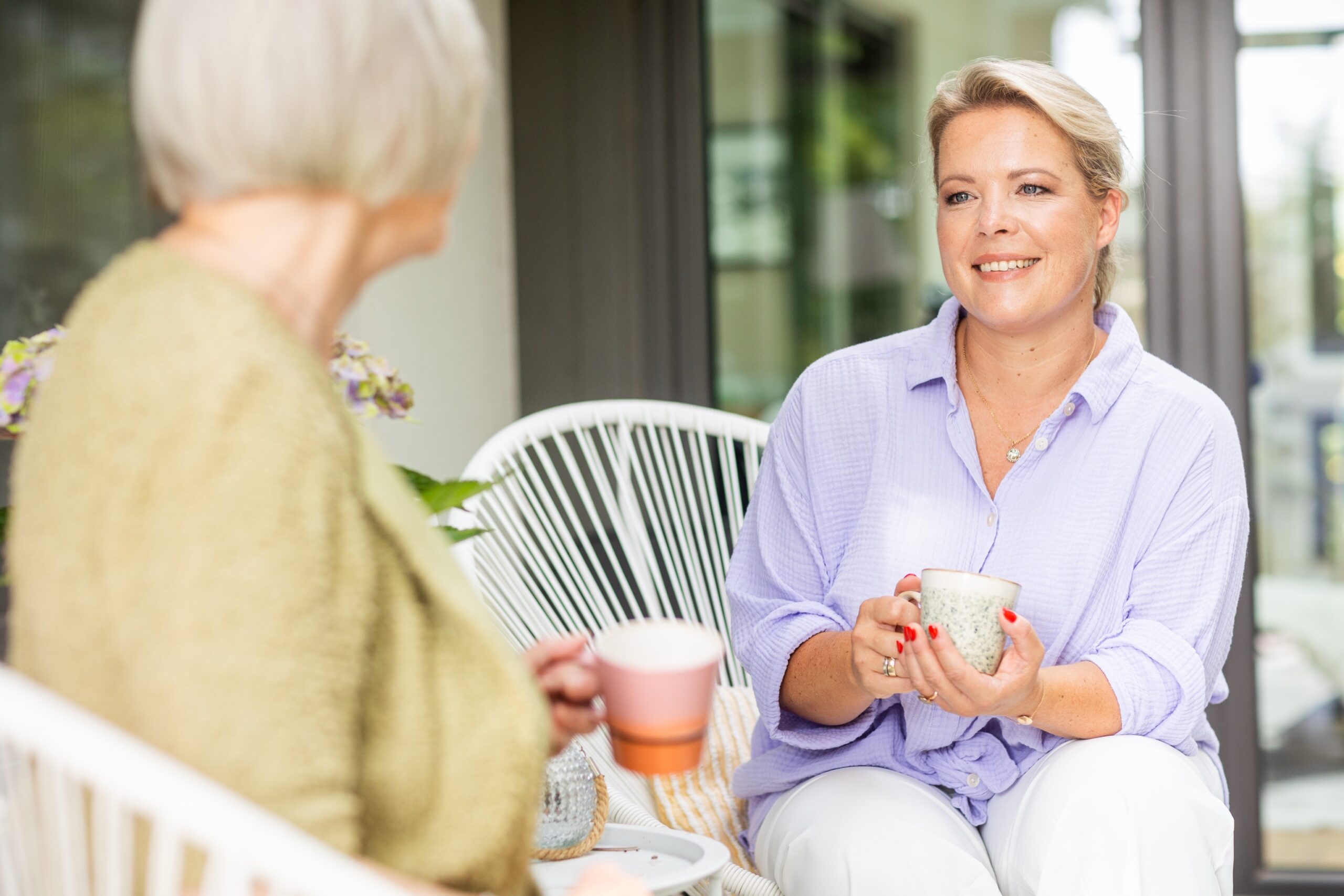 Alltagshelferin Linda Oebbeke sitzt mit einer älteren Frau auf einer Terrasse, beide halten eine Tasse in der Hand und unterhalten sich.